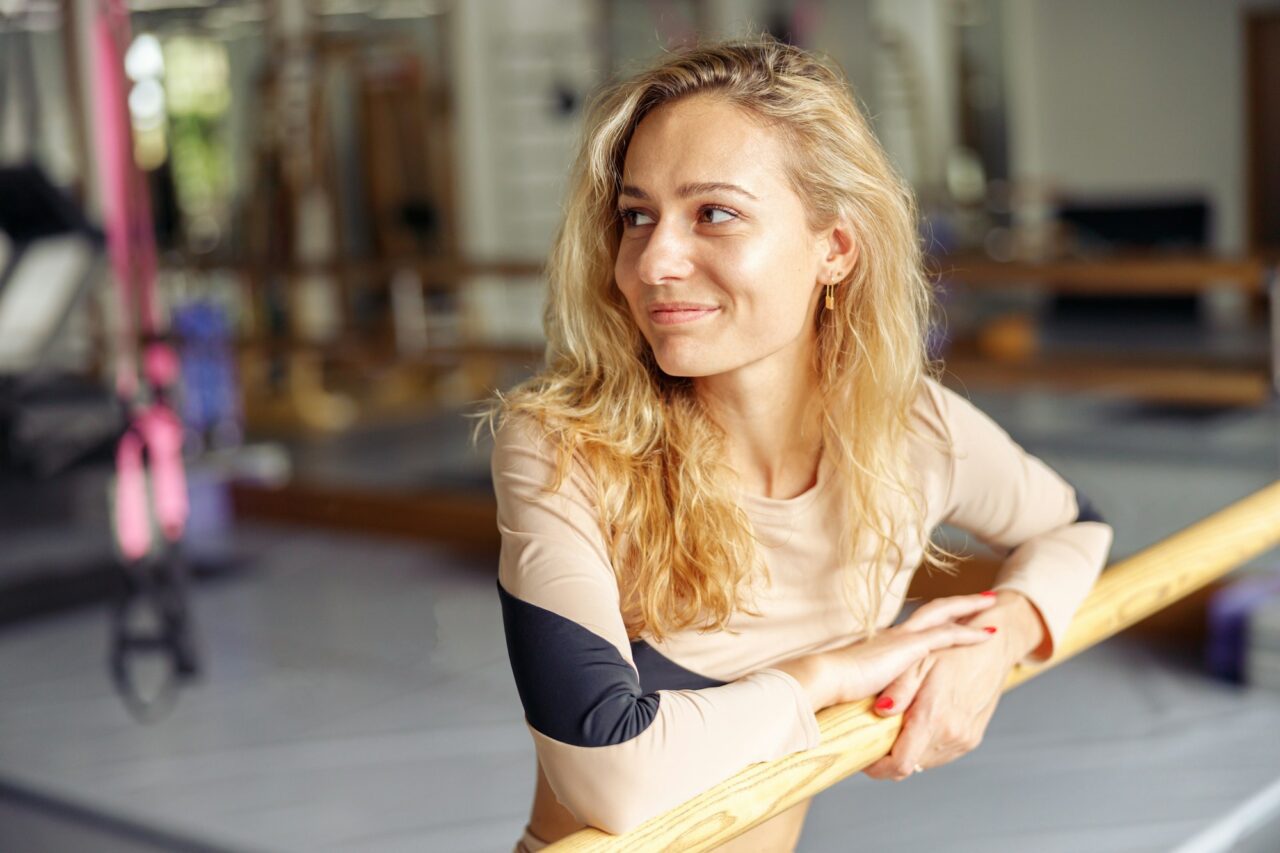 Poirtrait of attractive young woman, dancer standing near ballet barre in stretching studio