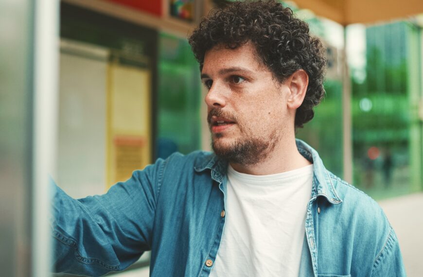 Close-up, young bearded man in denim shirt stands at bus stop and looks at the timetable
