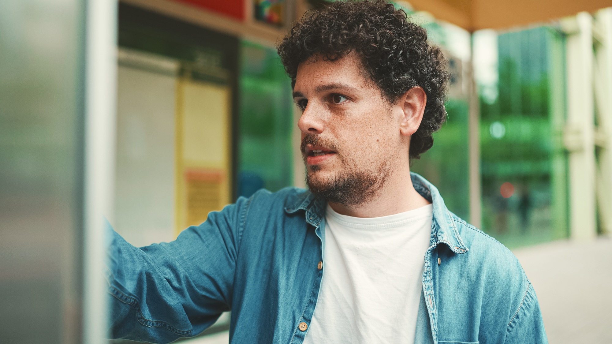 Close-up, young bearded man in denim shirt stands at bus stop and looks at the timetable