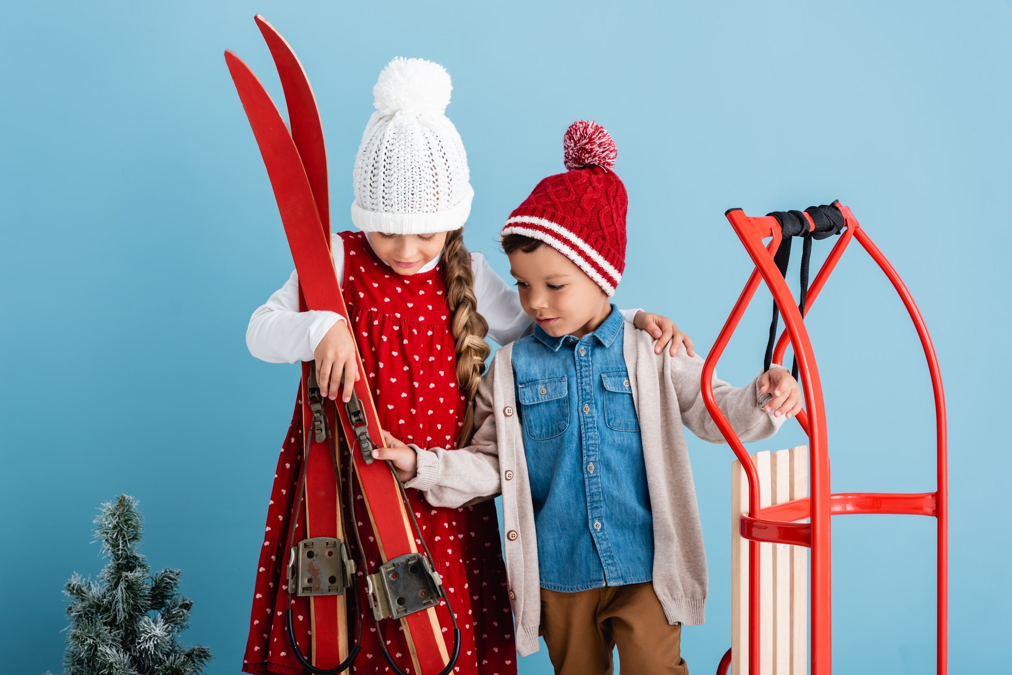 girl in winter outfit holding skis near brother with sleight on blue